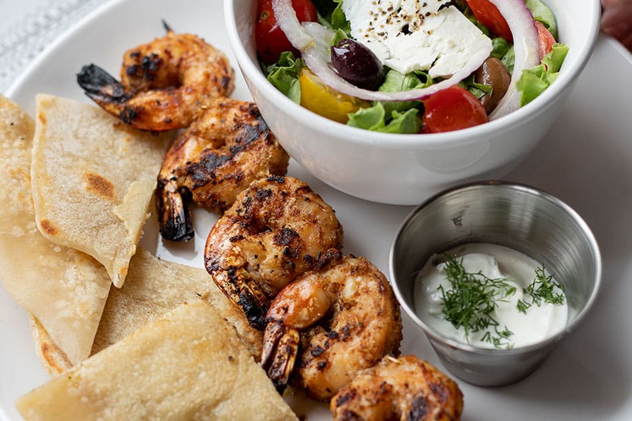 Marinated shrimp prepared and served on a skewer with a greek salad side and tzatziki sauce